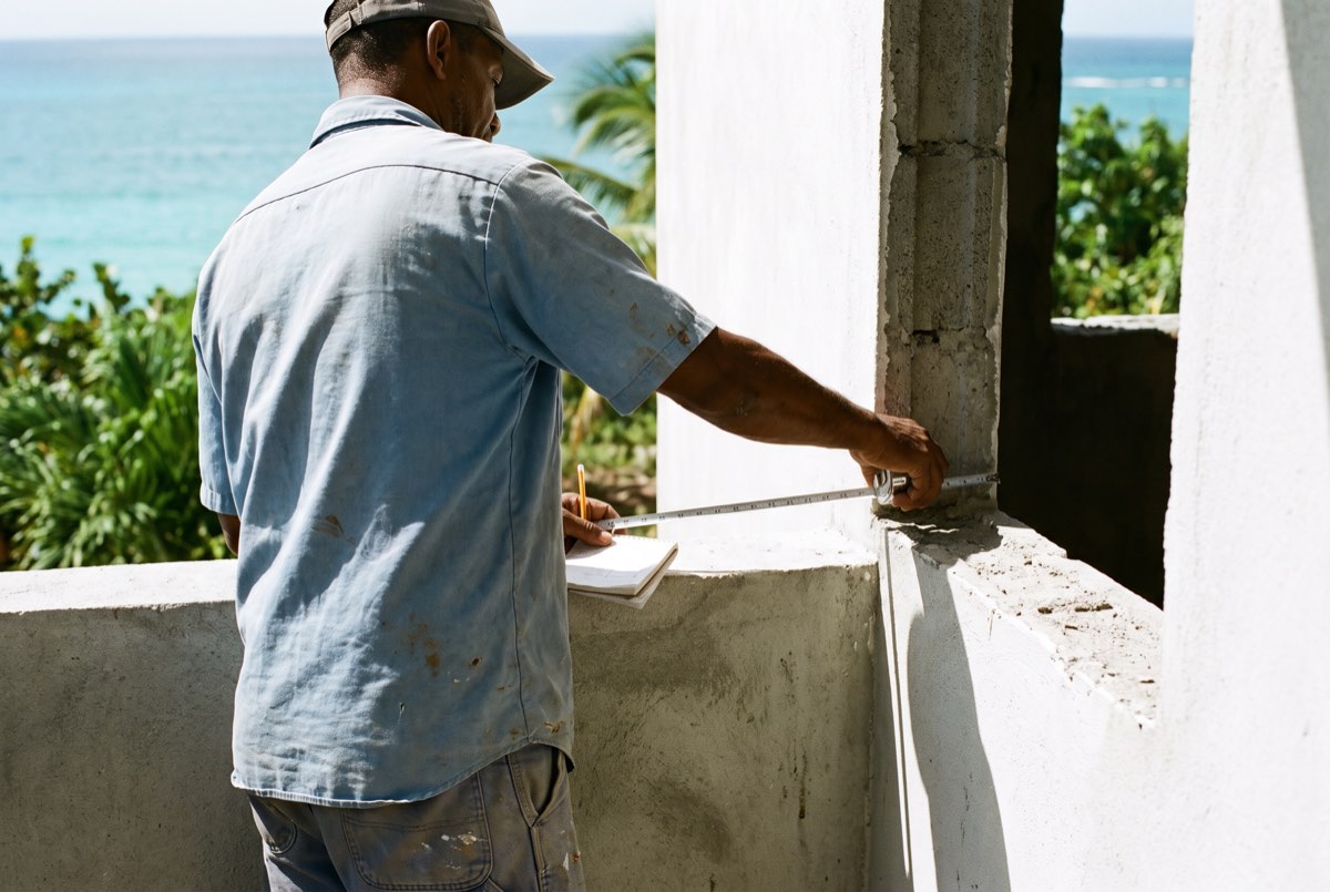 A tradesman measuring the width of a window opening on a Caribbean house terrace, notepad in hand.