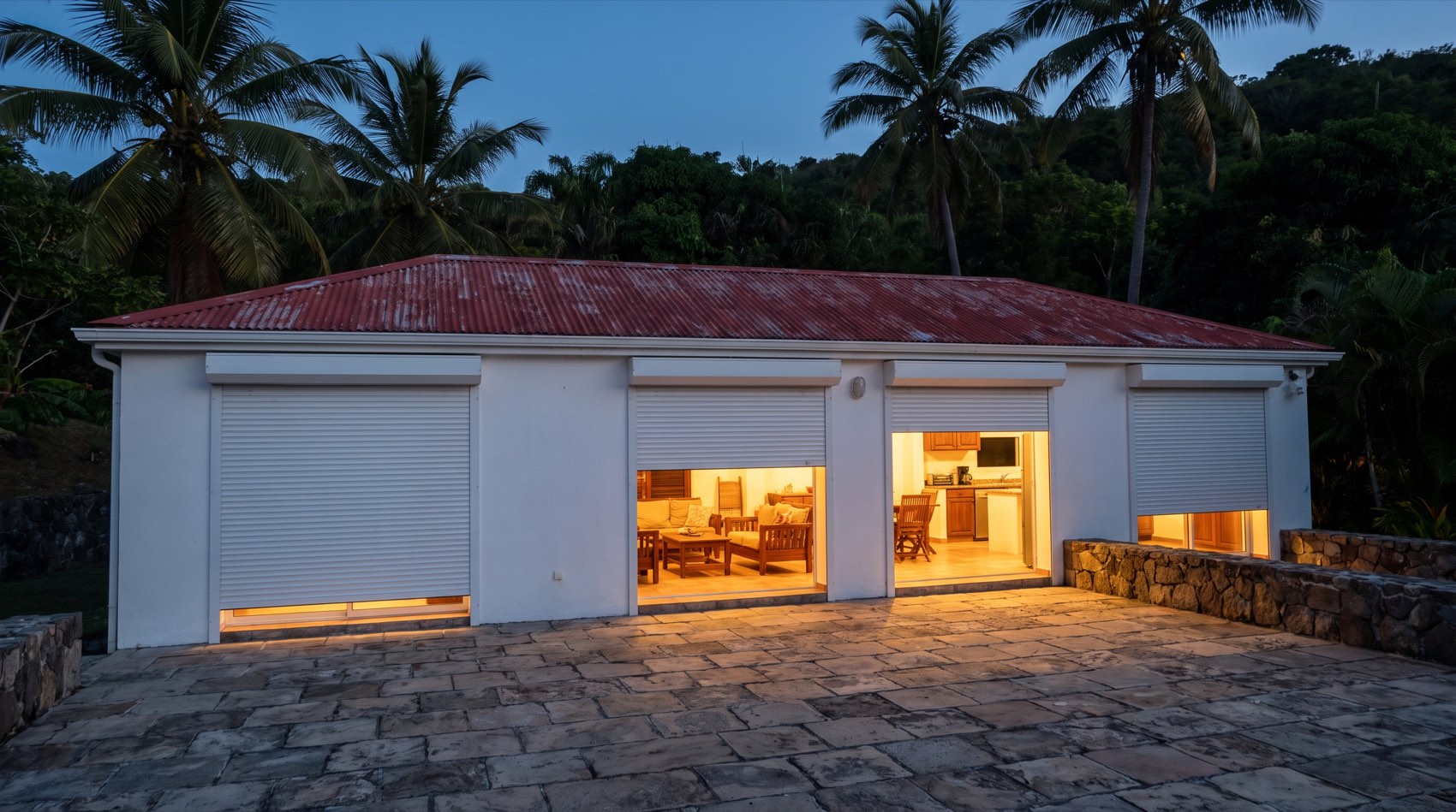 A white Caribbean villa at dusk with white roll shutters lowered to different heights across four wide openings, warm light spilling out beneath them.